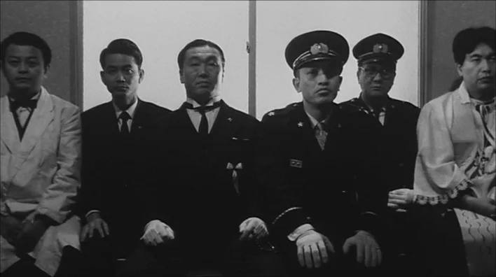 Death By Hanging (1968) movie still: medium — Six men in formal attire sit side by side in a bright, sparsely furnished room. They face the camera…