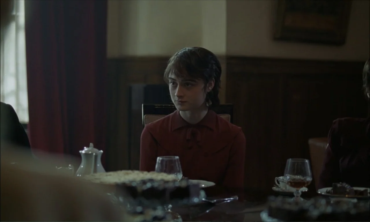 The Brutalist (2024) movie still: medium — A dimly lit room with wood-paneled walls and a set table. A young woman sits at the table looking toward…