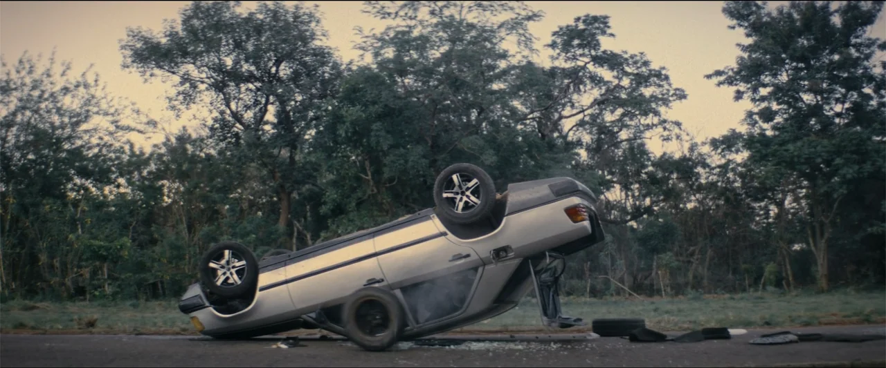 Ms. Kanyin (2025) movie still: wide — A silver car lies upside down on a paved road surrounded by dense trees. Smoke rises from the overturned…