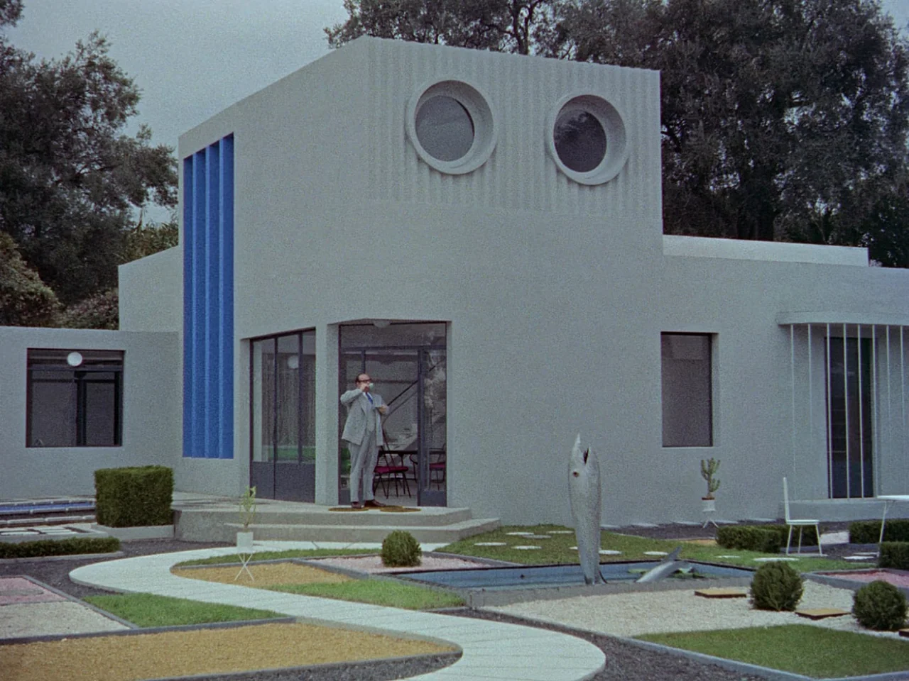 Mon Oncle (1958) movie still: wide — A minimalist grey villa features blue vertical accents and two large circular windows. A lone man stands in the…