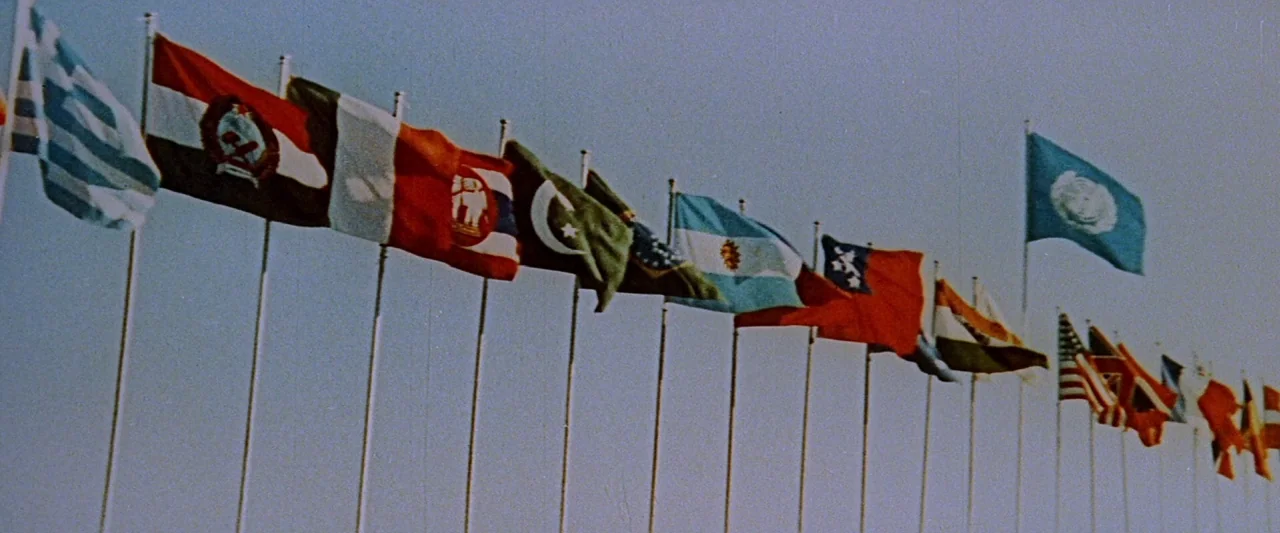 King Kong Vs. Godzilla (1962) movie still: wide — A long line of national flags flies on tall metal poles against a clear sky. The flags display…