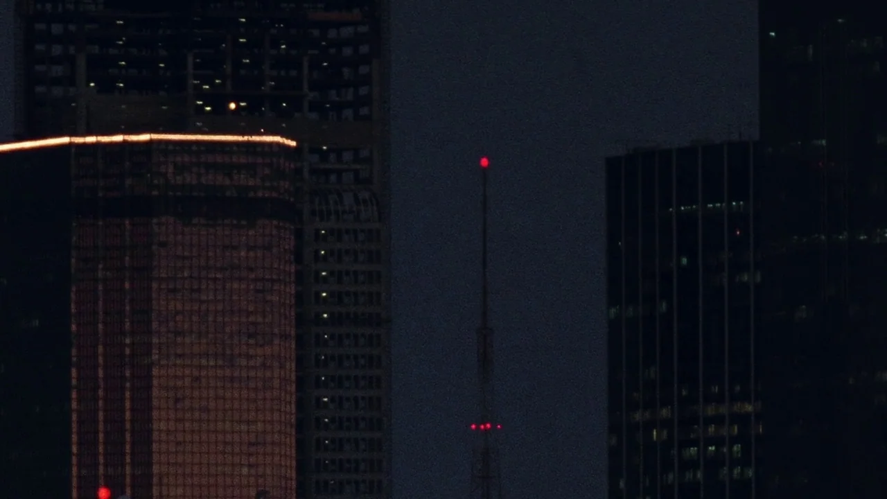 The Thin Blue Line (1988) movie still: establishing — A dark city skyline features tall glass skyscrapers under a night sky. A tall communications…