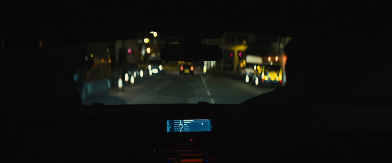 Locke (2013) movie still: wide — The view from inside a moving car at night shows a dimly lit road. Distant streetlights and headlights of other…