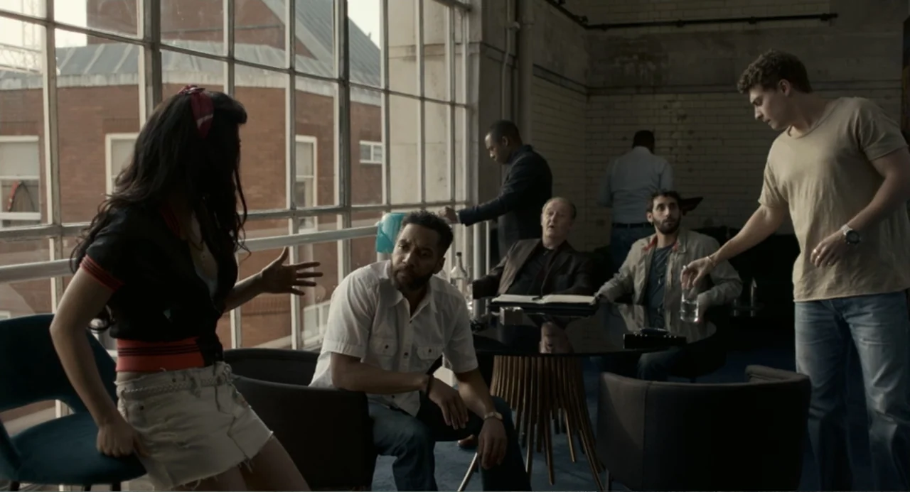 Back to Black (2024) movie still: medium — An industrial office space features large glass windows and white brick walls. A group of five people…