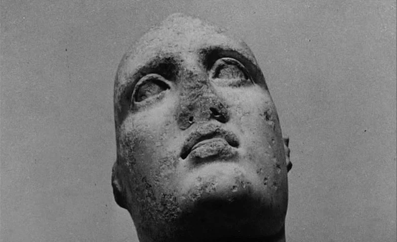 La Jetée (1962) movie still: closeup — A weathered stone sculpture head stands against a plain, light grey background. The sculpture is positioned…