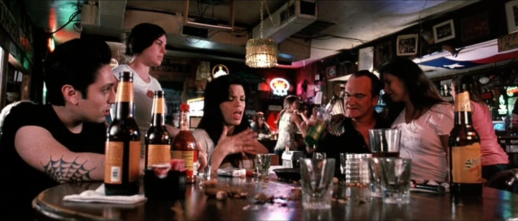 Death Proof movie still: medium — A dimly lit, wood-paneled dive bar interior with vintage decor and cluttered tables. A small group of friends sit…