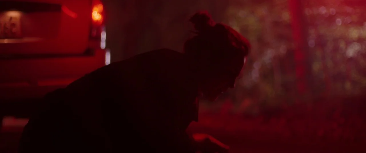 I Care A Lot (2020) movie still: medium — An outdoor area at night is illuminated by a pulsing red light. A person crouches low beside the rear…