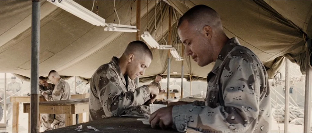 Jarhead movie still: medium — A military canvas tent contains rows of simple wooden tables and overhead fluorescent lights. Two soldiers in desert…