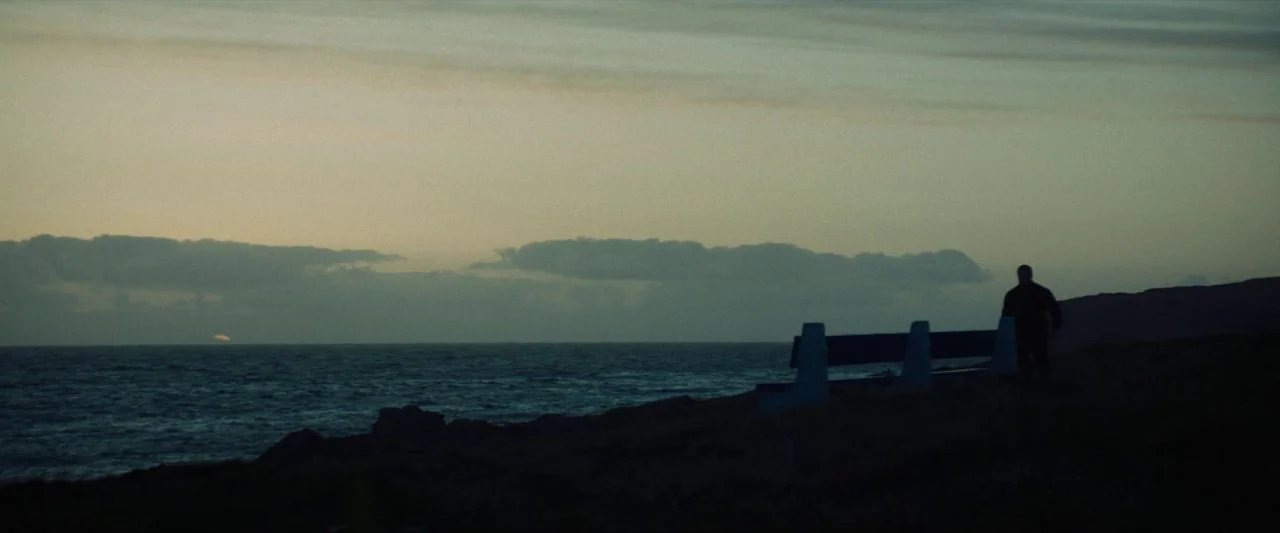 Calm With Horses (2019) movie still: wide — A rugged coastal cliffside overlooking the ocean at twilight with a single wooden bench. A lone figure…