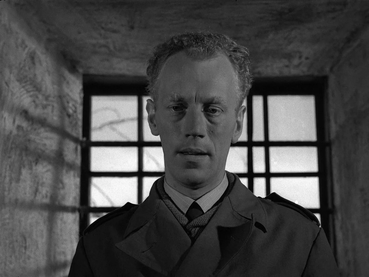 Winter Light (1963) movie still: closeup — A stone room features a window with iron bars set into the rear wall. A man in a collared shirt and coat…