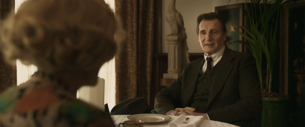 Marlowe (2023) movie still: medium — A dimly lit room with brown drapes and a decorative statue on a pedestal. A man sits at a dining table while…