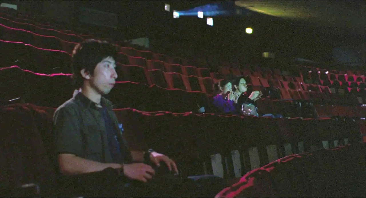 Goodbye, Dragon Inn (2003) movie still: medium — A dark movie theater with rows of empty red seats in the background. A young man sits in the…