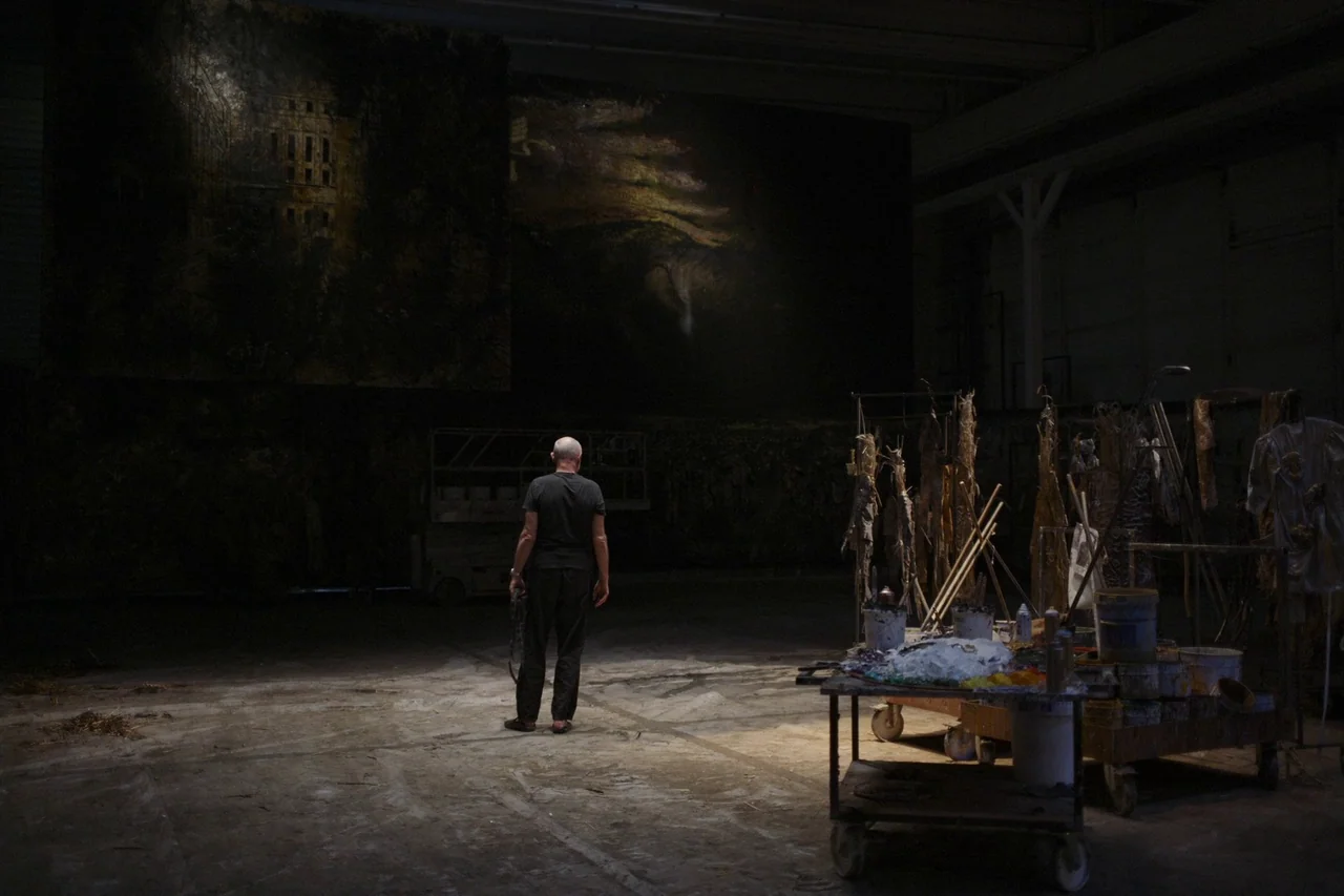 Anselm (2023) movie still: wide — A vast, dim studio space features large dark paintings mounted on the back wall. An elderly man stands in a…