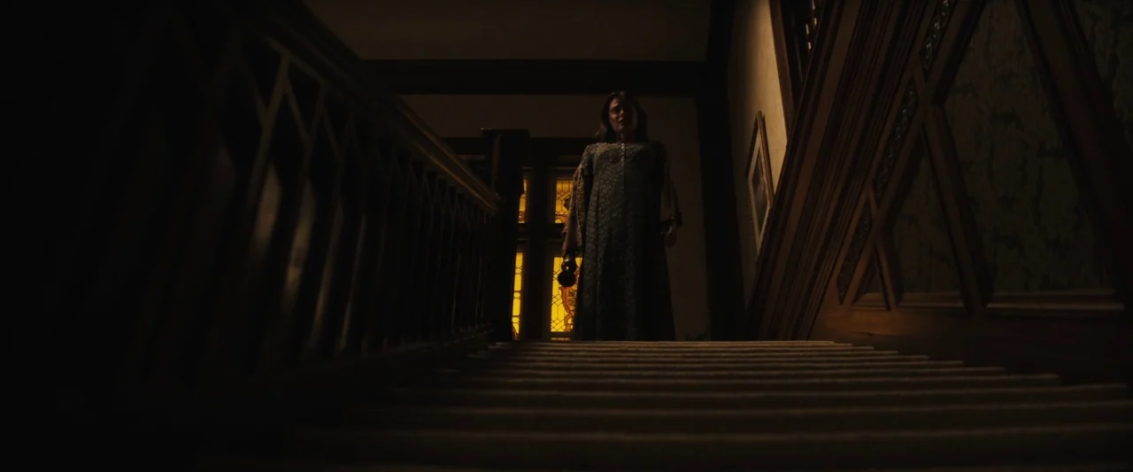 The Grudge (2020) movie still: fullbody — A dimly lit wooden staircase features a woman standing on the upper landing area. She holds a small lamp…