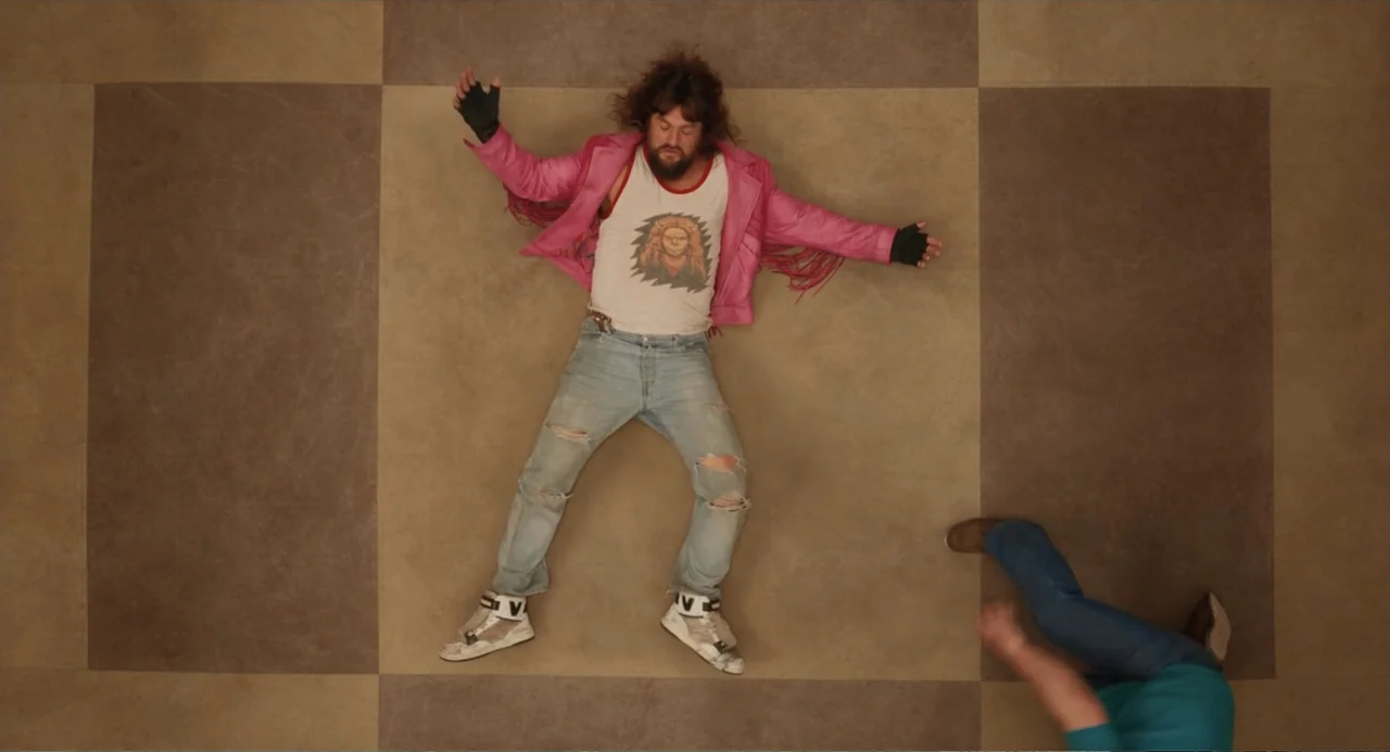 A Minecraft Movie (2025) movie still: fullbody — A square-tiled floor forms the background for two people in a large room. A man lies flat on his…