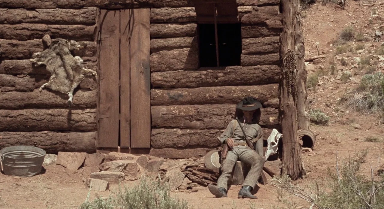 Ride In The Whirlwind (1966) movie still: fullbody — A rustic log structure sits in a dry, arid environment with red-brown dirt. A man in a cowboy…