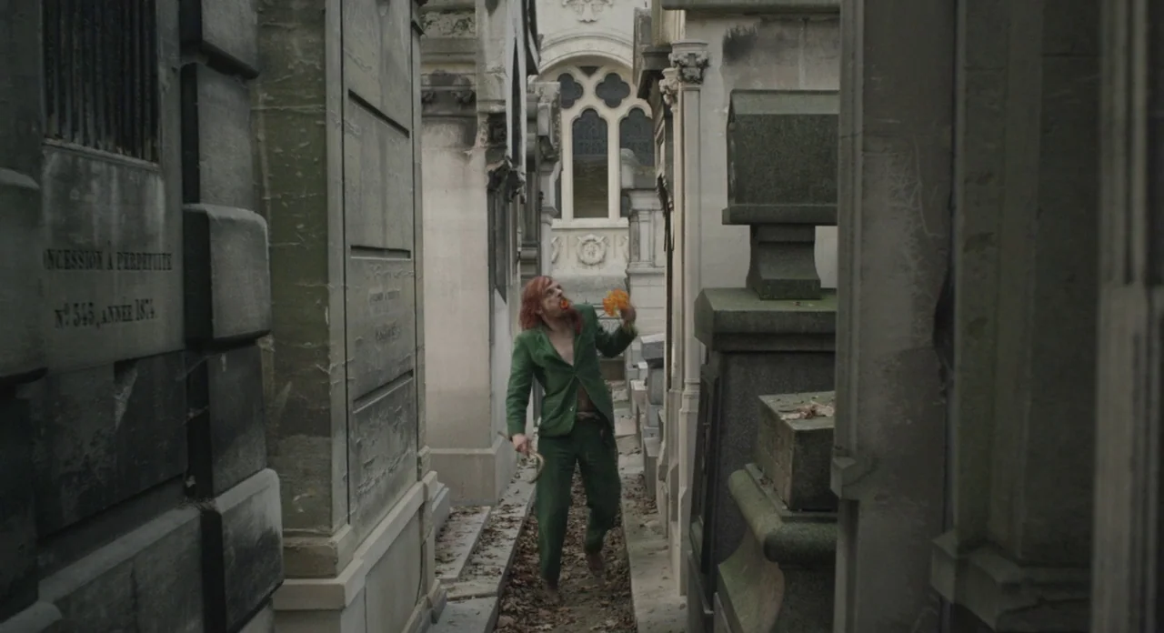Holy Motors (2012) movie still: fullbody — A narrow corridor flanked by tall, weathered stone mausoleums with arched architectural features. A man…