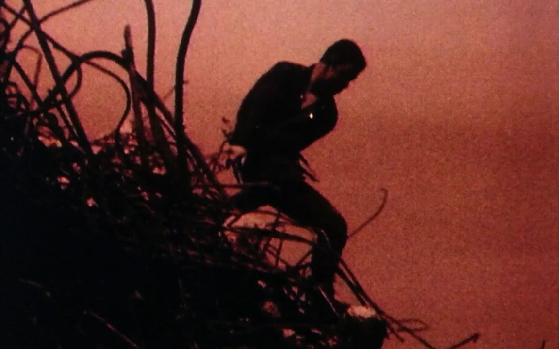 The Last of England (1987) movie still: fullbody — A pile of twisted metal debris sits against a stark, red-toned sky. A man climbs across the…