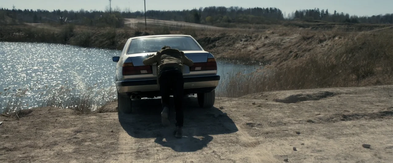 The Good Neighbor (2022) movie still: medium — A rural dirt path sits adjacent to a small pond under clear daylight. A man in a green jacket pushes…