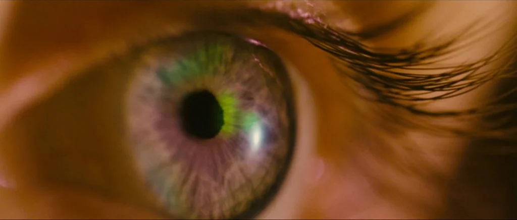 Sunshine movie still: closeup — A close-up view focuses on the details of a human eye. The iris displays purple and green hues reflecting internal…