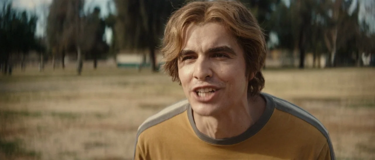 The Disaster Artist (2017) movie still: closeup — An open park with dry, yellow grass and blurred trees in the background. A young man with light…