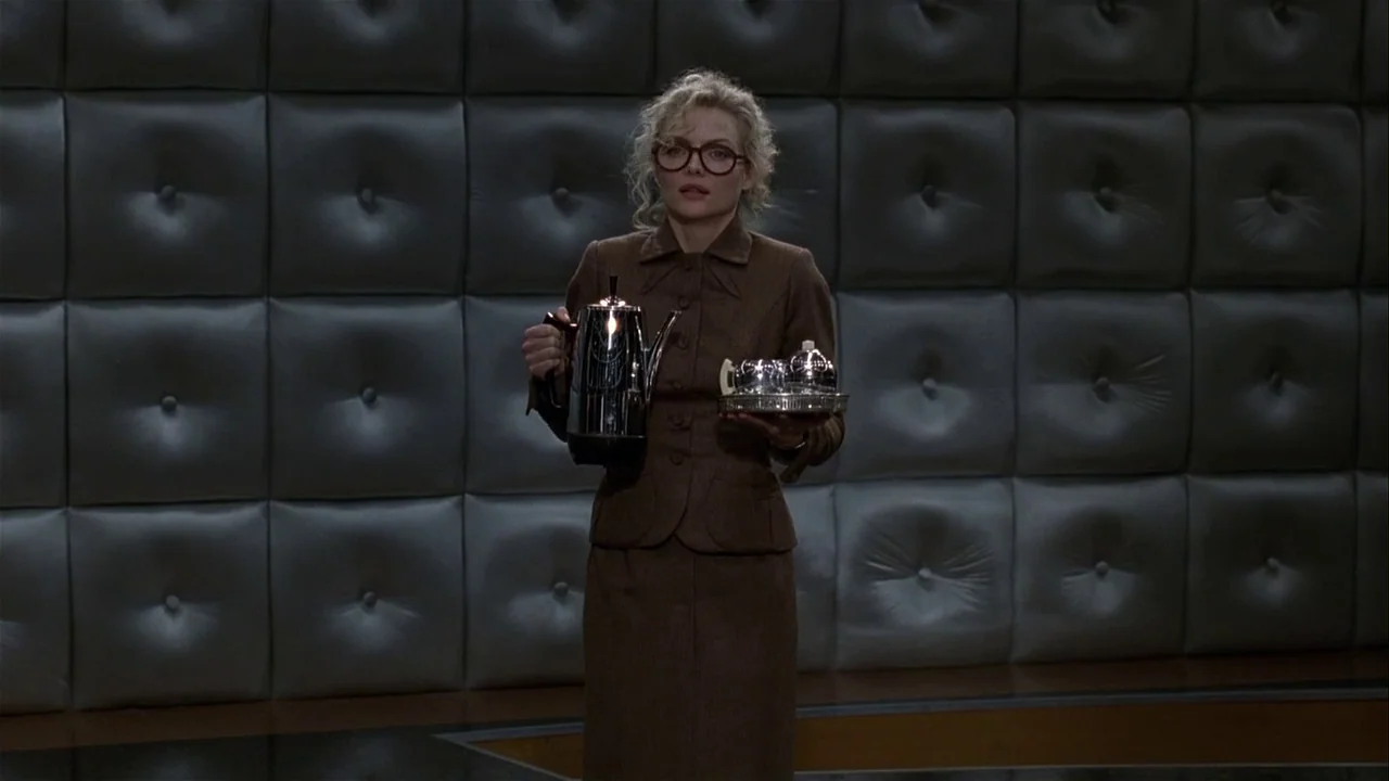 Batman Returns (1992) movie still: fullbody — A woman stands in front of a dark gray wall with square tufted padding. She holds a silver coffee pot…