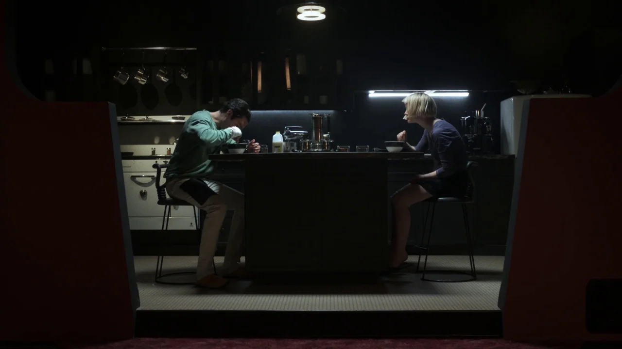 Piercing (2018) movie still: wide — A dark, modern dining area features two people sitting at a central table. The man and woman sit in profile…