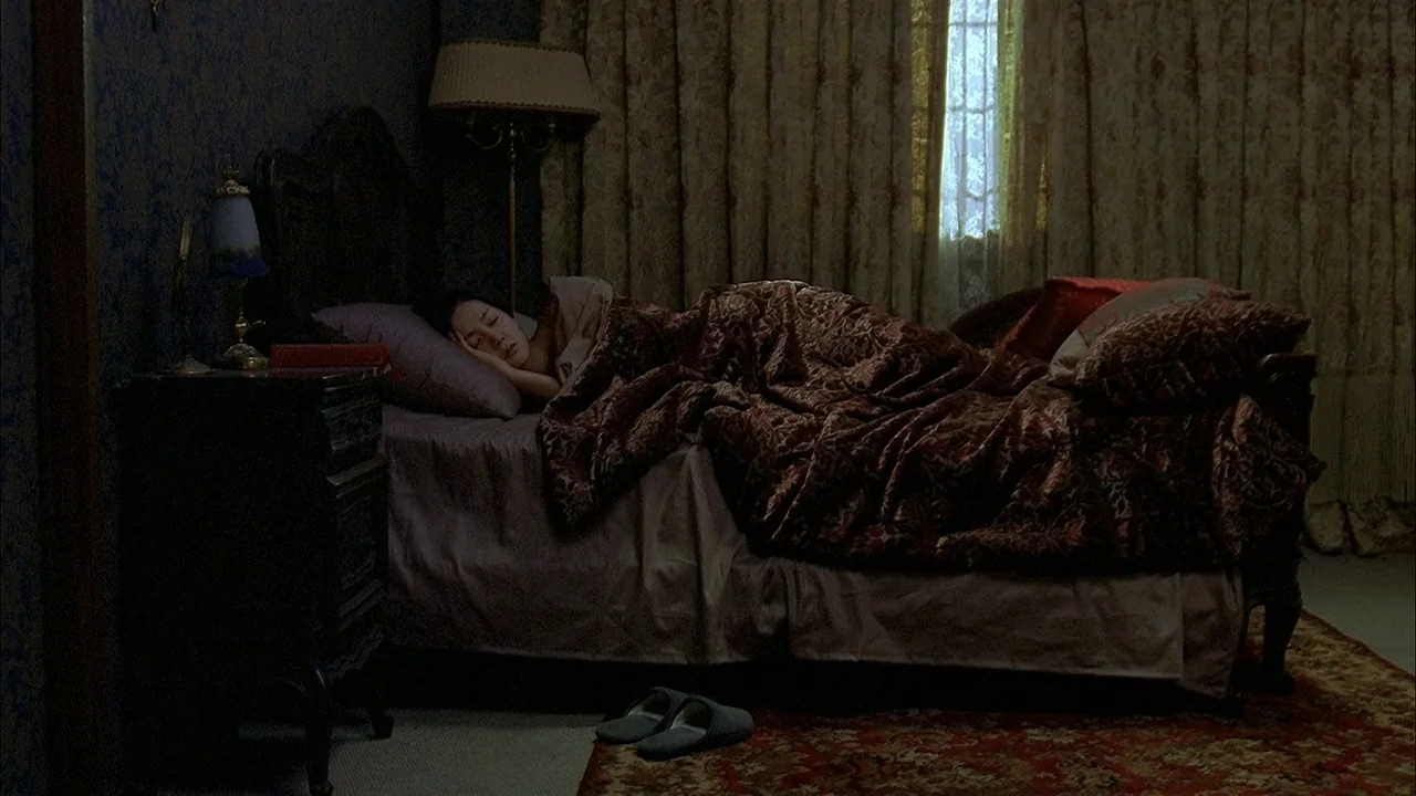 A Tale of Two Sisters (2003) movie still: wide — A dimly lit room with dark floral wallpaper features a heavy carved wooden bed. A woman sleeps…