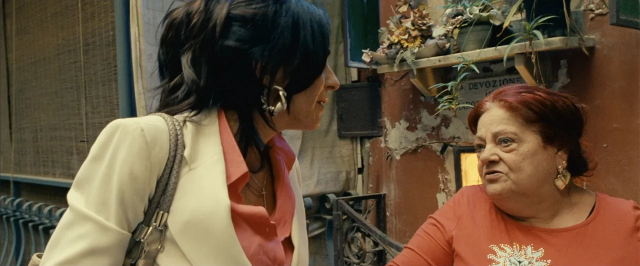 Reality (2023) movie still: medium — A narrow alleyway with terracotta-colored walls and a shelf of dried plants above. Two women stand side-by-side…