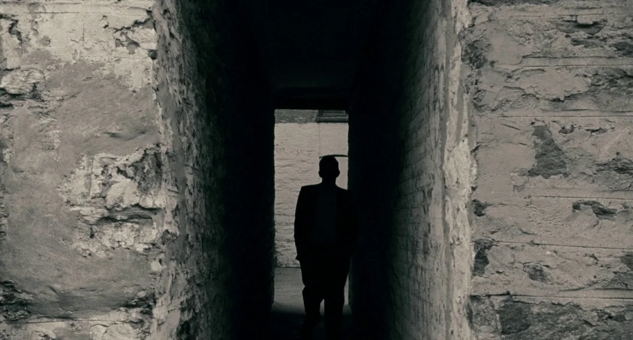 Howl (2010) movie still: wide — A narrow stone corridor features crumbling masonry on both sides leading to a bright exit. A lone figure in a suit…