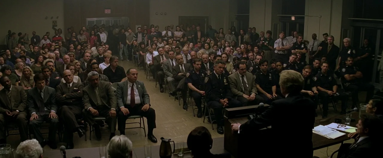 The Yards (2000) movie still: wide — A crowded auditorium filled with people sitting in metal chairs facing a podium. A man in a suit stands at the…