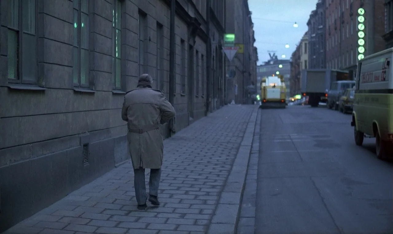 Man on the Roof (1976) movie still: fullbody — A stone-walled building lines a paved sidewalk in a city at dusk. An elderly man in a tan trench coat…