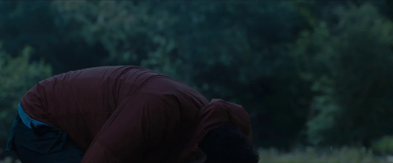 In The Earth (2021) movie still: medium — A dense forest environment at twilight with dark, blurred foliage in the background. A person wearing a…
