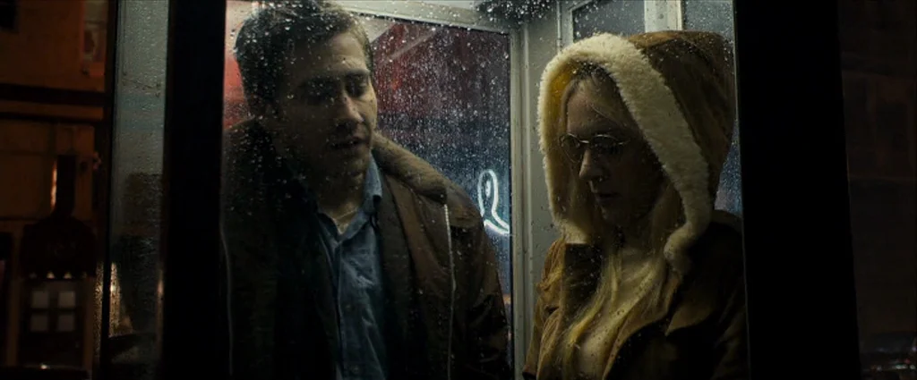 Zodiac (2007) movie still: medium — A rain-streaked glass phone booth at night contains a man and a woman. They stand together while rain droplets…
