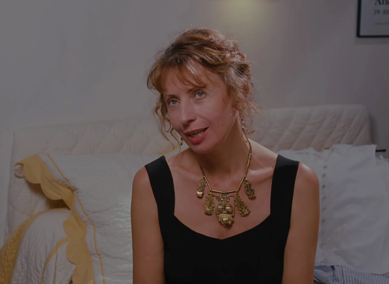 Autumn Tale (1998) movie still: medium — A woman sits in front of a white quilted bedspread with yellow trim. She is positioned centered in the…