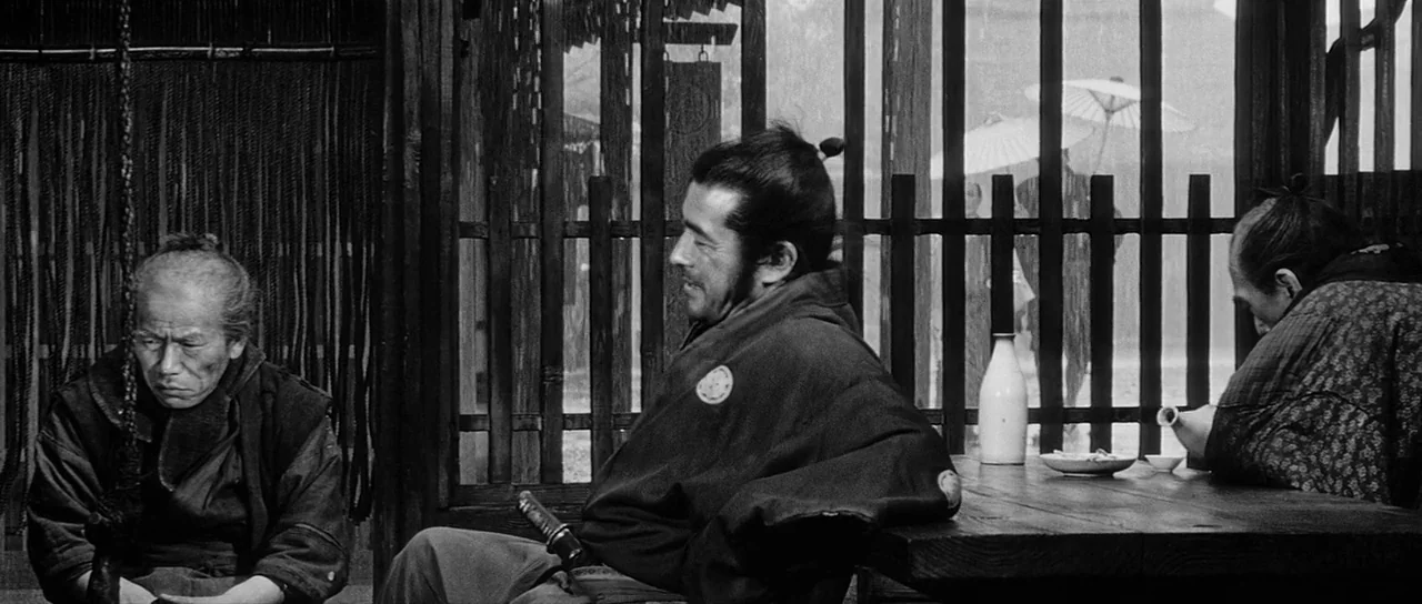 Yojimbo (1961) movie still: medium — A wooden shelter features slatted walls looking out onto a rainy street scene. Three men sit at a low wooden…