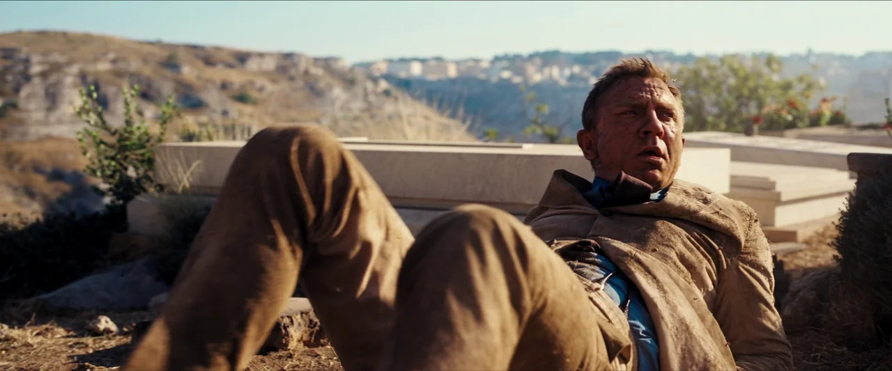 No Time To Die (2021) movie still: medium — An arid outdoor burial ground with white stone monuments under a bright sky. A man lies on the dirt…