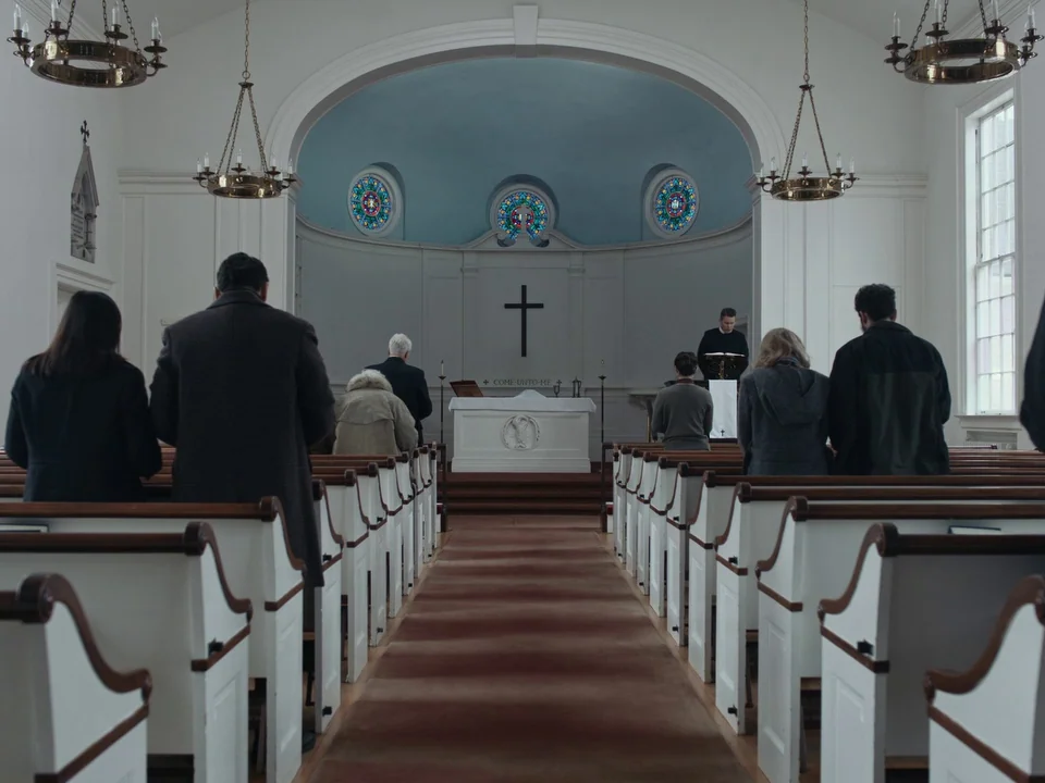 First Reformed (2017) movie still: wide — A white-walled, symmetrical church interior features rows of wooden pews and a central altar. Several…