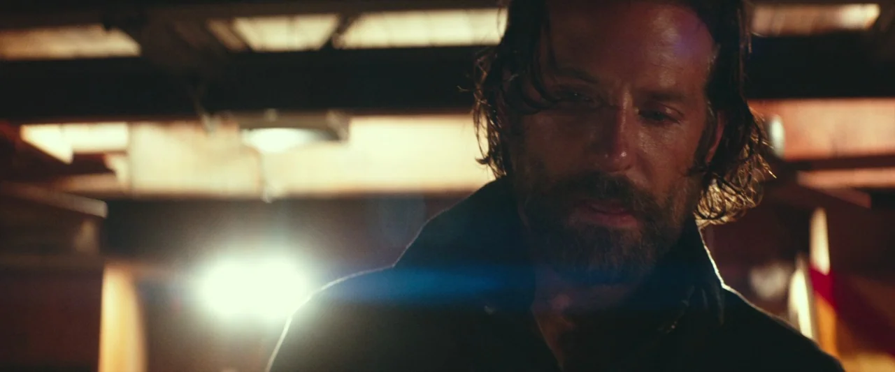 A Star Is Born (2018) movie still: closeup — A dimly lit industrial space features exposed wooden rafters and a blurred work light. A bearded man…