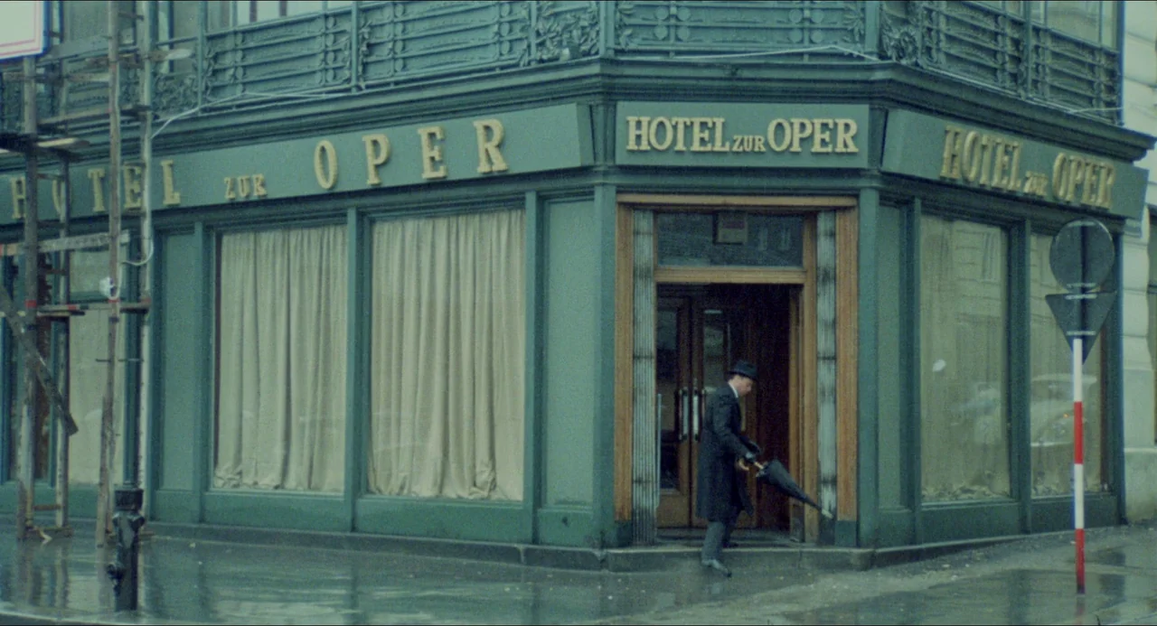 The Night Porter (1974) movie still: wide — A corner building features green painted paneling and large windows with cream curtains. A man in a dark…