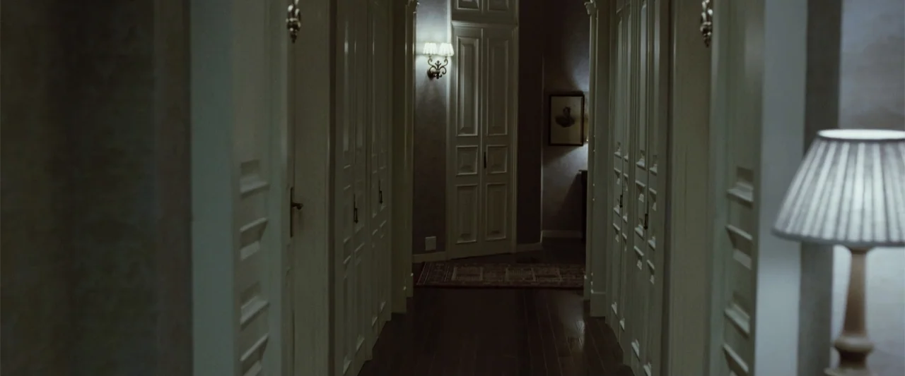Anna (2013) movie still: wide — A dimly lit narrow residential hallway features white paneled doors and dark hardwood floors. A wall sconce and a…