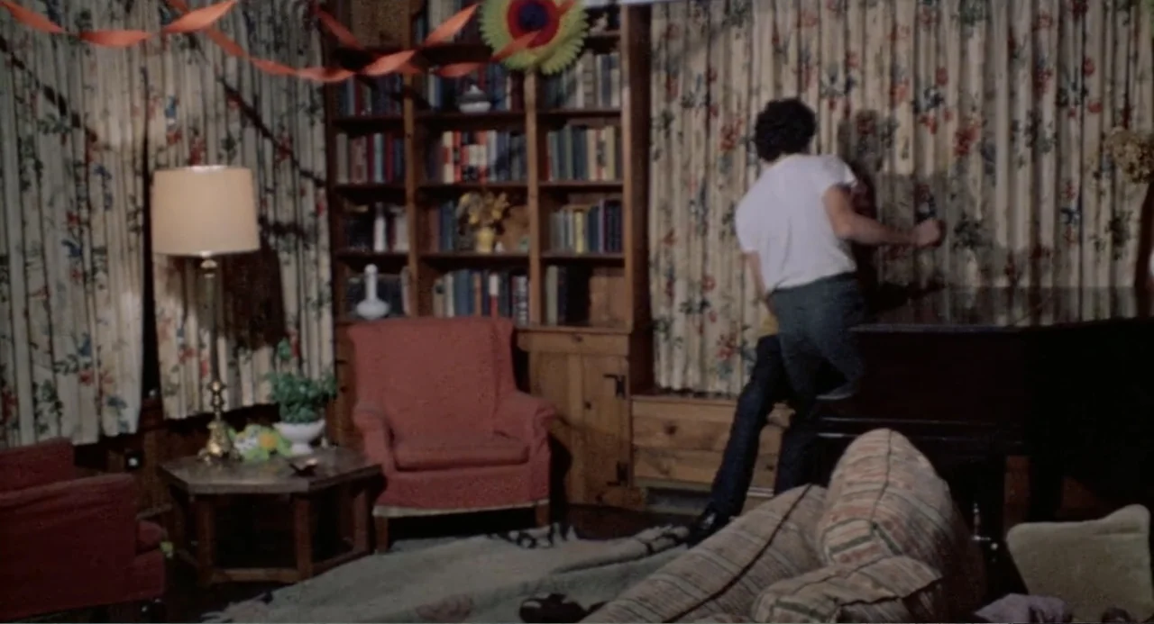 The Last House on the Left (2009) movie still: medium — A 1970s living room features a wooden bookshelf, floral curtains, and red armchairs. Two…