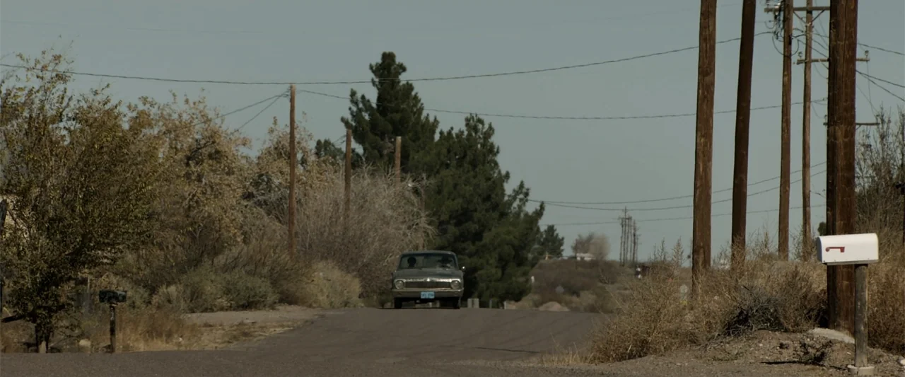 Cry Macho (2021) movie still: wide — An asphalt road cuts through a dry, sparsely vegetated rural desert landscape. A vintage sedan drives down the…