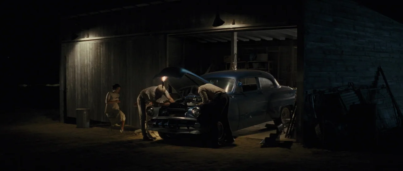 Loving (2016) movie still: wide — A dark, wooden structure housing a vintage blue vehicle at night. Two men examine the engine under a work light…
