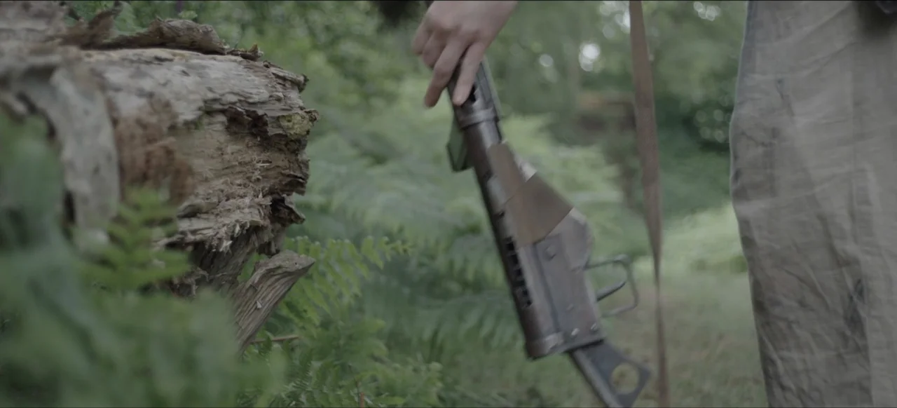 We Go In at Dawn (2020) movie still: medium — A person stands in a dense area filled with ferns and a fallen tree. The individual holds a metallic…