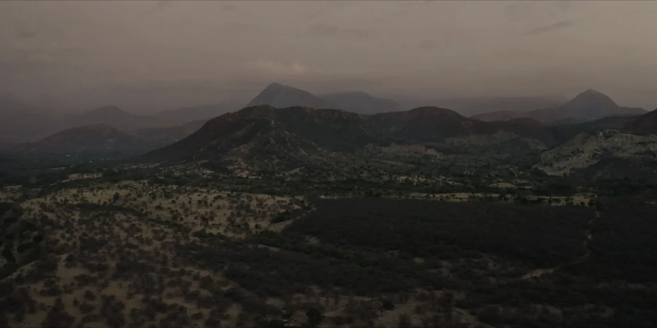 Leyenda Viva (2022) movie still: establishing — A expansive mountain range under a thick layer of grey evening clouds. Rolling hills and dark shrubs…