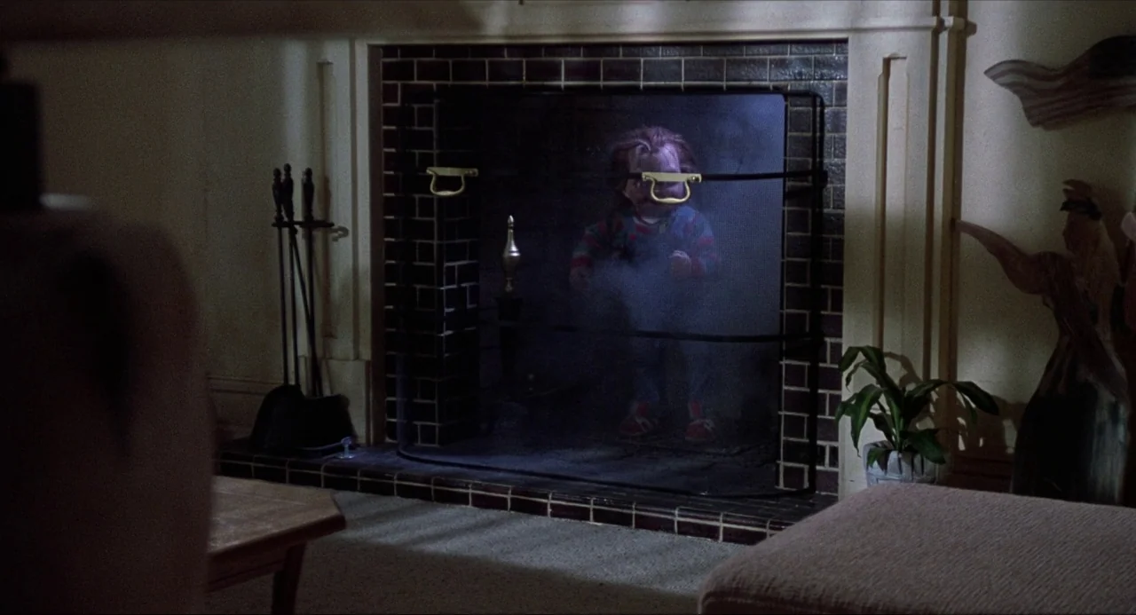 Child’s Play (1988) movie still: medium — A dark, wood-paneled room features a large brick fireplace with a fine mesh screen. A small doll stands…