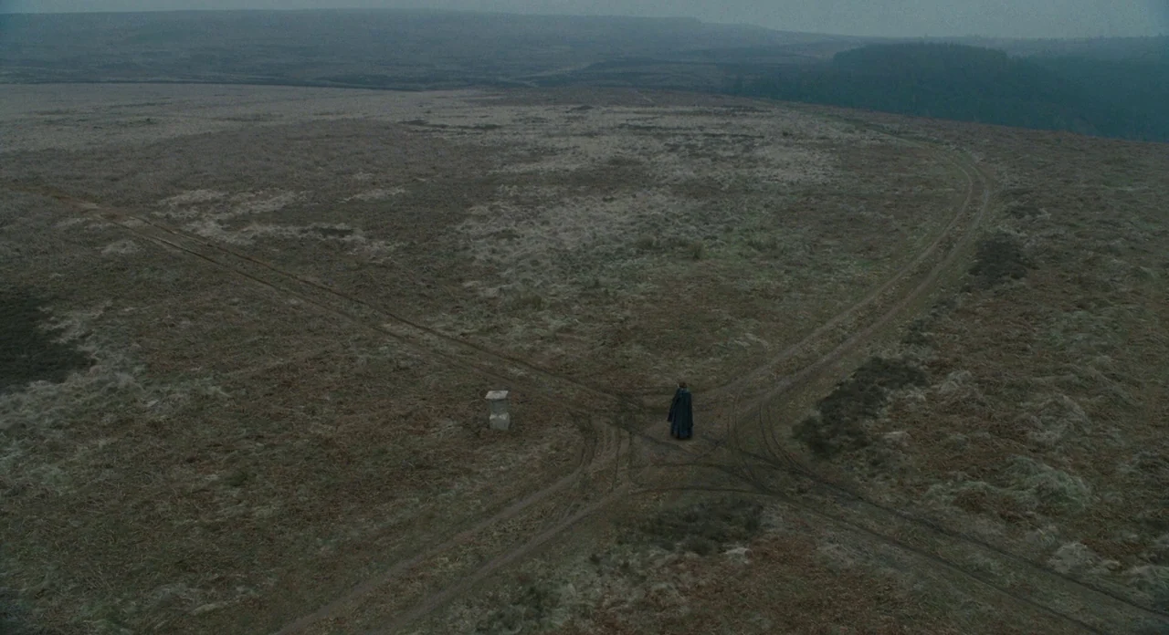 Jane Eyre (2011) movie still: wide — A vast, desolate moorland landscape features intersecting dirt paths under a grey, overcast sky. A lone figure…