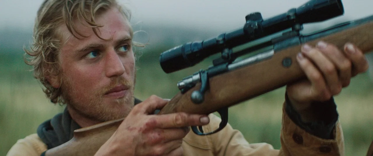 Beast (2017) movie still: medium — An open rural landscape with blurred greenery in the background. A young man aims a scoped hunting rifle forward…