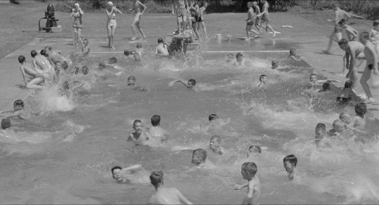 A Face In The Crowd (1957) movie still: wide — A crowded outdoor concrete swimming pool is packed with dozens of children playing together. The…